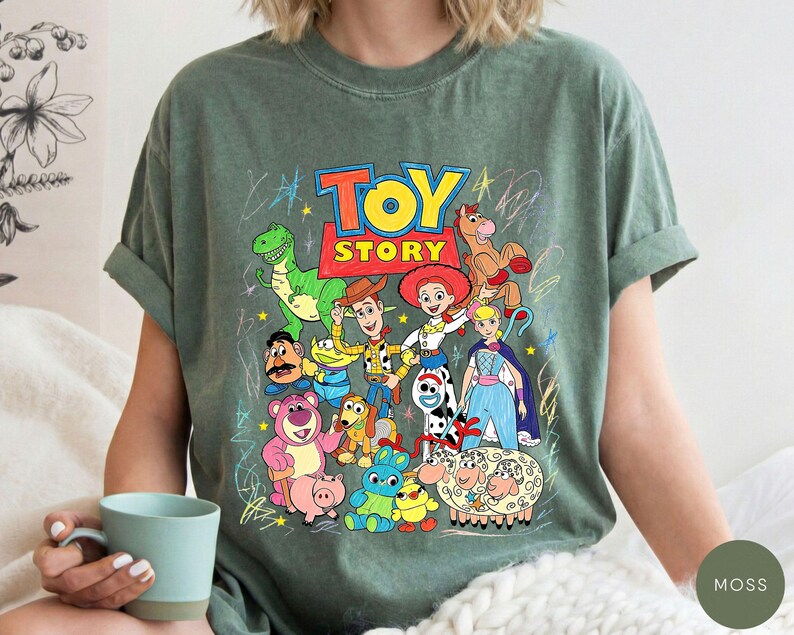 May include: A moss green short-sleeve t-shirt featuring a colourful graphic print of Toy Story characters including Woody, Jessie, Buzz Lightyear, Rex, and others. The design includes the Toy Story logo and scribbled star and line accents.
