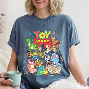 May include: A blue jean coloured t-shirt featuring a colourful graphic print of Toy Story characters including Woody, Buzz Lightyear, Jessie, Rex, and others. The design includes the Toy Story logo and scribbled star and line accents.