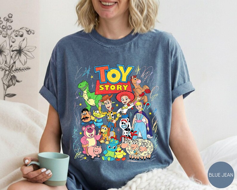May include: A blue jean coloured t-shirt featuring a colourful graphic print of Toy Story characters including Woody, Buzz Lightyear, Jessie, Rex, and others. The design includes the Toy Story logo and scribbled star and line accents.