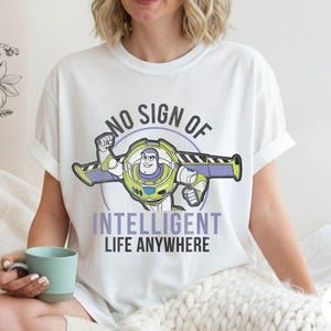 Puede incluir: Camiseta blanca con un dibujo animado de Buzz Lightyear y el texto "NO SIGN OF INTELLIGENT LIFE ANYWHERE". El dise&ntilde;o presenta a Buzz con su traje espacial con detalles en morado y verde.