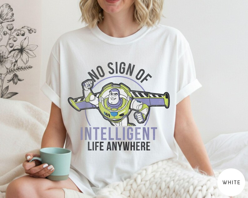 Puede incluir: Camiseta blanca con un dibujo animado de Buzz Lightyear y el texto "NO SIGN OF INTELLIGENT LIFE ANYWHERE". El dise&ntilde;o presenta a Buzz con su traje espacial con detalles en morado y verde.