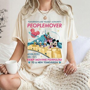 May include: Ivory-colored t-shirt featuring a colorful graphic of the Tomorrowland Transit Authority PeopleMover ride with Mickey Mouse, Minnie Mouse, Donald Duck, and Goofy. The shirt includes text that reads "PEOPLEMOVER" and "KEEP MOVING FORWARD".