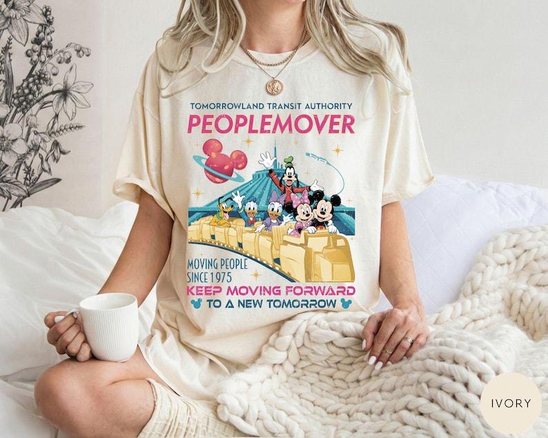 May include: Ivory-colored t-shirt featuring a colorful graphic of the Tomorrowland Transit Authority PeopleMover ride with Mickey Mouse, Minnie Mouse, Donald Duck, and Goofy. The shirt includes text that reads "PEOPLEMOVER" and "KEEP MOVING FORWARD".