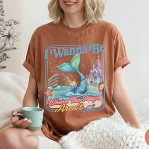 May include: A brown t-shirt featuring a mermaid graphic with the text "I Wanna Be Where The People Aren't." The design includes a mermaid, castle, and ocean elements in a retro style. The shirt has short sleeves and a relaxed fit.