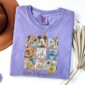 Może przedstawiać: Fioletowa koszulka Comfort Colors z siatką postaci Disneya, w tym Myszki Miki, Myszki Minnie i Goofy'ego. Tekst "Walt Disney World" znajduje się na dole. Widoczny jest r&oacute;wnież złoty naszyjnik.