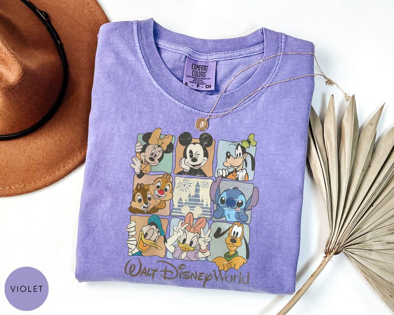 Może przedstawiać: Fioletowa koszulka Comfort Colors z siatką postaci Disneya, w tym Myszki Miki, Myszki Minnie i Goofy'ego. Tekst "Walt Disney World" znajduje się na dole. Widoczny jest r&oacute;wnież złoty naszyjnik.
