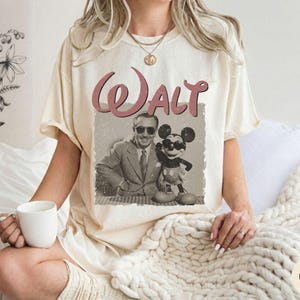 Puede incluir: Camiseta color marfil con un gr&aacute;fico vintage de Walt Disney y Mickey Mouse. La palabra "Walt" est&aacute; impresa en una fuente cursiva roja encima de la imagen. La camiseta tiene un corte holgado y mangas cortas.