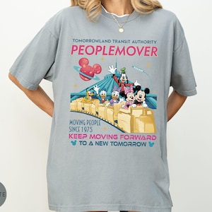 May include: A light gray t-shirt featuring the Tomorrowland Transit Authority Peoplemover ride with Disney characters. The design includes the text "MOVING PEOPLE SINCE 1975" and "KEEP MOVING FORWARD TO A NEW TOMORROW".