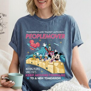 May include: Blue jean colored t-shirt featuring a graphic of Disney characters on a PeopleMover ride. The shirt includes the text "TOMORROWLAND TRANSIT AUTHORITY PEOPLEMOVER" and "MOVING PEOPLE SINCE 1975".