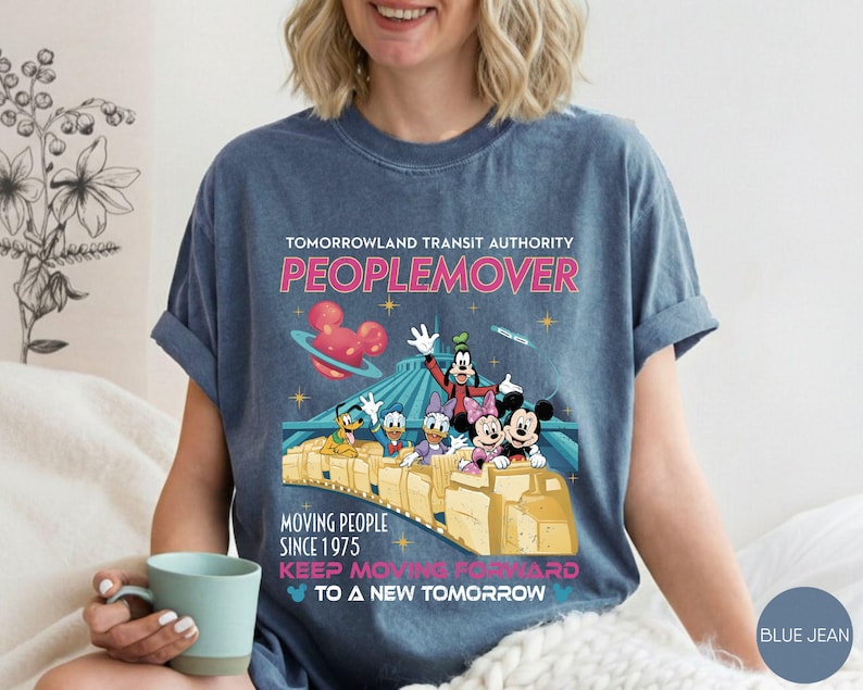 May include: Blue jean colored t-shirt featuring a graphic of Disney characters on a PeopleMover ride. The shirt includes the text "TOMORROWLAND TRANSIT AUTHORITY PEOPLEMOVER" and "MOVING PEOPLE SINCE 1975".