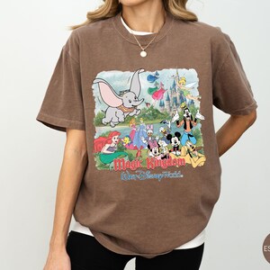 May include: A brown short-sleeve t-shirt featuring a graphic print of various Disney characters including Dumbo, Mickey Mouse, and Ariel in front of a castle. The text reads Magic Kingdom and Walt Disney World in red and blue lettering on the front.