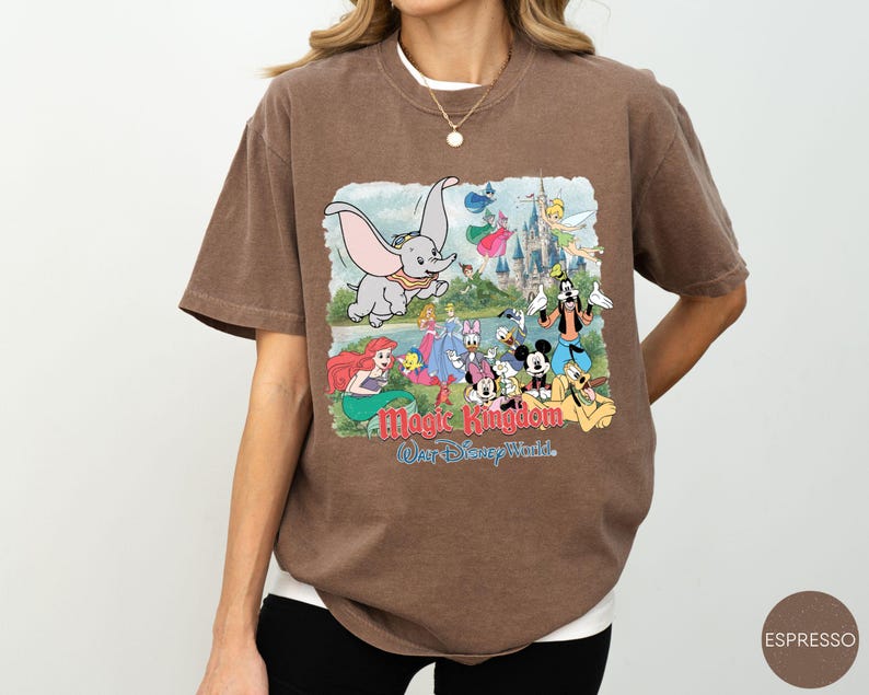 May include: A brown short-sleeve t-shirt featuring a graphic print of various Disney characters including Dumbo, Mickey Mouse, and Ariel in front of a castle. The text reads Magic Kingdom and Walt Disney World in red and blue lettering on the front.