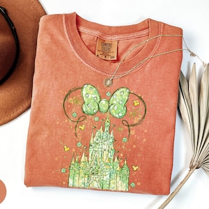 K&ouml;nnte beinhalten: Korallenfarbenes T-Shirt mit einem gr&uuml;n-wei&szlig;en Schleifendesign und einer Schlossgrafik. Das T-Shirt hat einen Rundhalsausschnitt. Eine goldene Halskette liegt &uuml;ber dem Shirt. Ein brauner Hut und eine getrocknete Pflanze sind im Hintergrund.