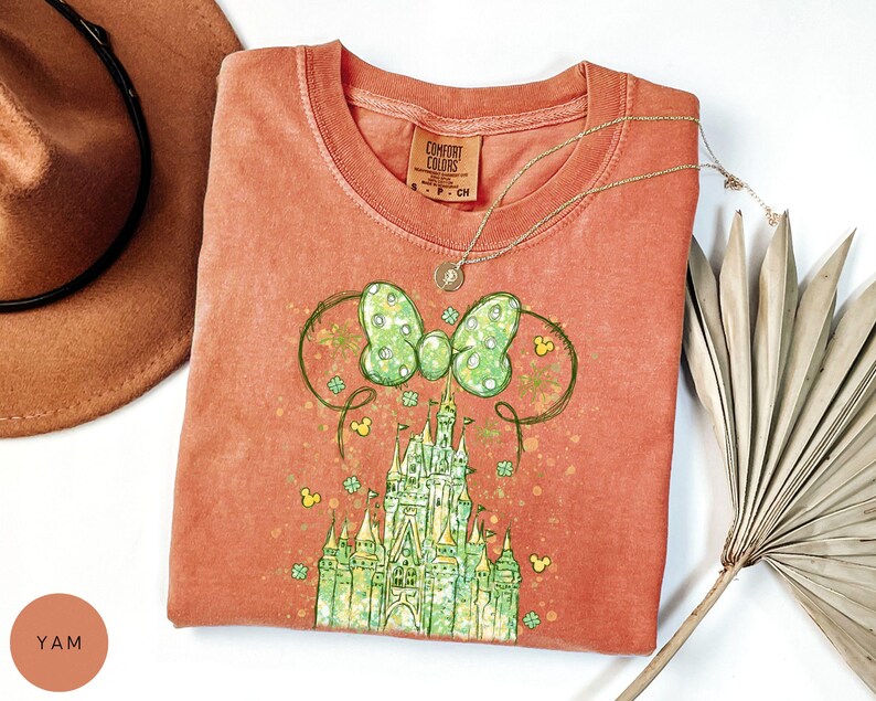 K&ouml;nnte beinhalten: Korallenfarbenes T-Shirt mit einem gr&uuml;n-wei&szlig;en Schleifendesign und einer Schlossgrafik. Das T-Shirt hat einen Rundhalsausschnitt. Eine goldene Halskette liegt &uuml;ber dem Shirt. Ein brauner Hut und eine getrocknete Pflanze sind im Hintergrund.