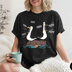 May include: Black t-shirt with a cartoon design of two white seagulls with yellow beaks and feet, each saying "MOM!" above a rock in the water. A small orange fish is in the water. The shirt is a comfortable fit.