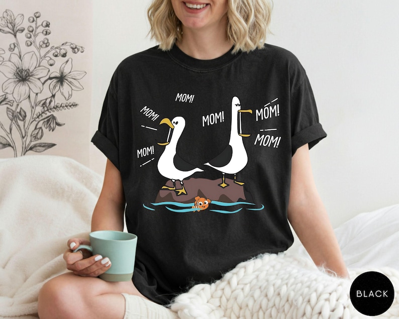 May include: Black t-shirt with a cartoon design of two white seagulls with yellow beaks and feet, each saying "MOM!" above a rock in the water. A small orange fish is in the water. The shirt is a comfortable fit.