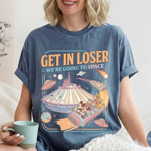 Pode incluir: Camiseta azul jeans com um design gr&aacute;fico retr&ocirc;. O design apresenta o texto "GET IN LOSER WE'RE GOING TO SPACE" com uma ilustra&ccedil;&atilde;o colorida de um passeio em um parque tem&aacute;tico espacial e personagens.