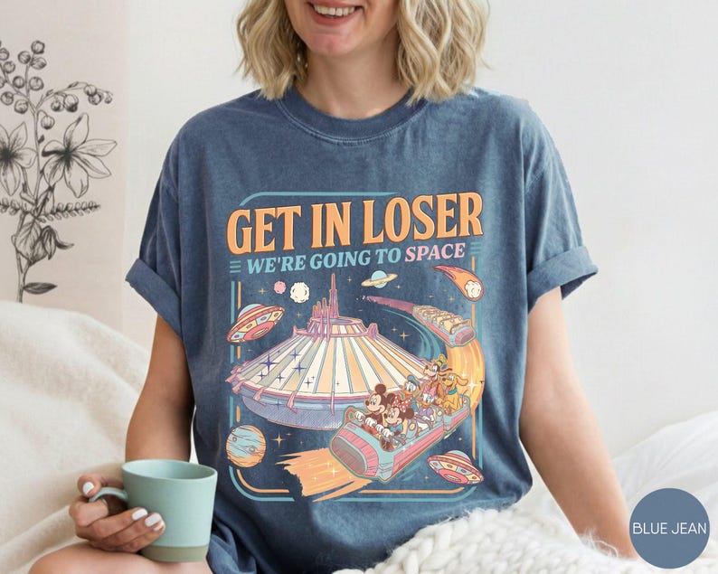 Pode incluir: Camiseta azul jeans com um design gr&aacute;fico retr&ocirc;. O design apresenta o texto "GET IN LOSER WE'RE GOING TO SPACE" com uma ilustra&ccedil;&atilde;o colorida de um passeio em um parque tem&aacute;tico espacial e personagens.
