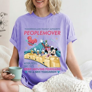 May include: A violet-colored t-shirt featuring a graphic of the Tomorrowland Transit Authority PeopleMover ride with Mickey Mouse, Minnie Mouse, Goofy, Donald Duck, and Daisy Duck. The shirt has the text "MOVING PEOPLE SINCE 1975" and "KEEP MOVING FORWARD TO A NEW TOMORROW".