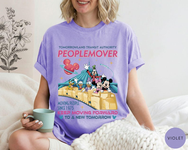 May include: A violet-colored t-shirt featuring a graphic of the Tomorrowland Transit Authority PeopleMover ride with Mickey Mouse, Minnie Mouse, Goofy, Donald Duck, and Daisy Duck. The shirt has the text "MOVING PEOPLE SINCE 1975" and "KEEP MOVING FORWARD TO A NEW TOMORROW".