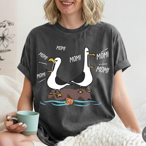 May include: Dark grey t-shirt with a cartoon design of two seagulls on a rock, with the word "MOM!" in speech bubbles. A small orange fish swims in the water below. The t-shirt has a relaxed fit.