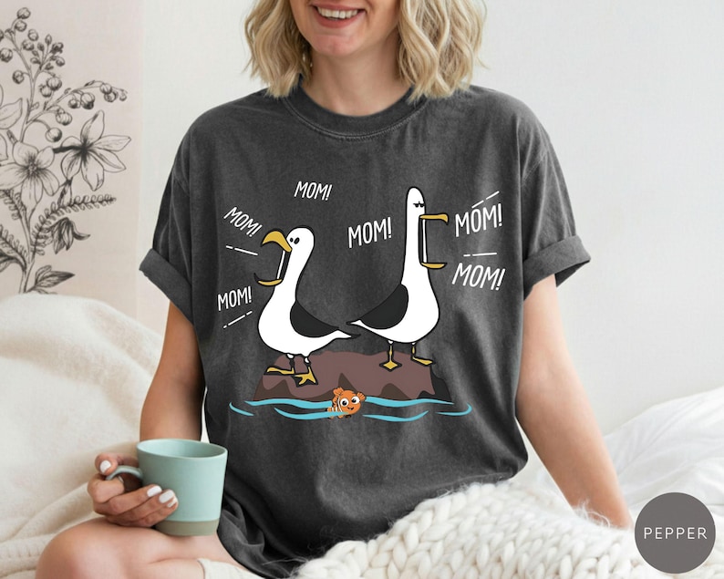 May include: Dark grey t-shirt with a cartoon design of two seagulls on a rock, with the word "MOM!" in speech bubbles. A small orange fish swims in the water below. The t-shirt has a relaxed fit.