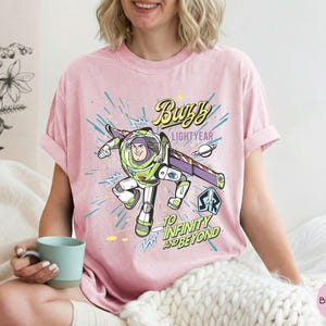 Peut inclure: T-shirt rose clair avec un graphisme de Buzz l'&Eacute;clair, le texte "Buzz Lightyear" et "To Infinity and Beyond". Le design comprend un th&egrave;me spatial avec une plan&egrave;te et des &eacute;toiles.