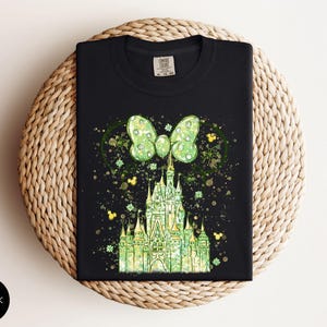 K&ouml;nnte beinhalten: Schwarzes T-Shirt mit einem gr&uuml;nen Schlossdesign, einer Schleife und Mickey Mouse-Silhouetten. Das Design hat einen Aquarelleffekt mit goldenen und gr&uuml;nen Akzenten. Das T-Shirt ist auf einer gewebten Oberfl&auml;che gefaltet.