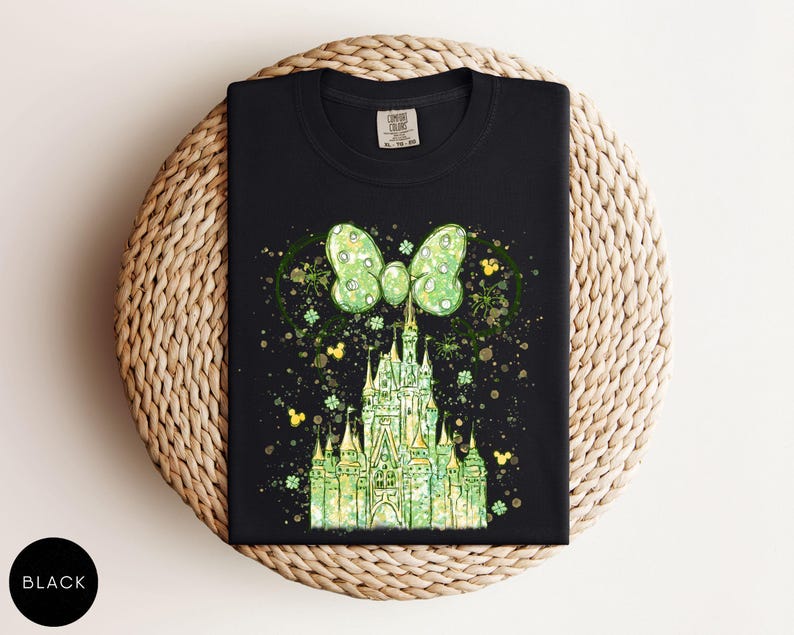 K&ouml;nnte beinhalten: Schwarzes T-Shirt mit einem gr&uuml;nen Schlossdesign, einer Schleife und Mickey Mouse-Silhouetten. Das Design hat einen Aquarelleffekt mit goldenen und gr&uuml;nen Akzenten. Das T-Shirt ist auf einer gewebten Oberfl&auml;che gefaltet.