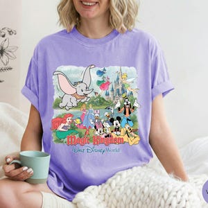 May include: A light purple short-sleeve t-shirt featuring a graphic print of various Disney characters including Dumbo, Ariel, Mickey Mouse, and Goofy in front of a castle. The text Magic Kingdom Walt Disney World is printed below the characters.