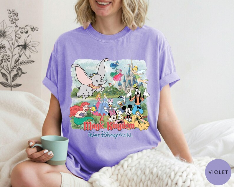 May include: A light purple short-sleeve t-shirt featuring a graphic print of various Disney characters including Dumbo, Ariel, Mickey Mouse, and Goofy in front of a castle. The text Magic Kingdom Walt Disney World is printed below the characters.