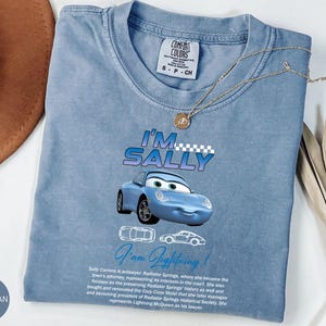 Puede incluir: Camiseta azul vaquero con una imagen de dibujos animados de Sally Carrera de la pel&iacute;cula Cars. La camiseta tiene el texto "I'm Sally" y "I'm Lightning!" impreso. Un collar de oro est&aacute; drapeado sobre la camiseta.