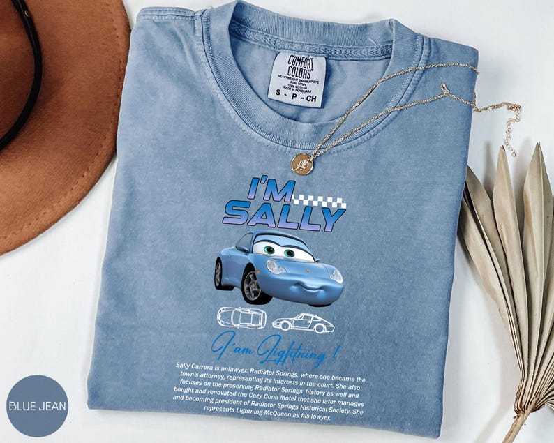 Puede incluir: Camiseta azul vaquero con una imagen de dibujos animados de Sally Carrera de la pel&iacute;cula Cars. La camiseta tiene el texto "I'm Sally" y "I'm Lightning!" impreso. Un collar de oro est&aacute; drapeado sobre la camiseta.