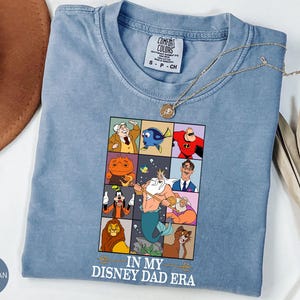 May include: Blue jean Comfort Colors t-shirt with a graphic featuring Disney characters in a grid. The graphic includes characters like Mr. Incredible, Dory, and King Triton. The text "IN MY DISNEY DAD ERA" is printed below the grid.
