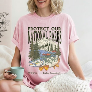 May include: Pink t-shirt with a graphic that says "Protect Our National Parks." The graphic features a mountain range, trees, a lake, and a fish. The text "Explore Responsibly" is also included.