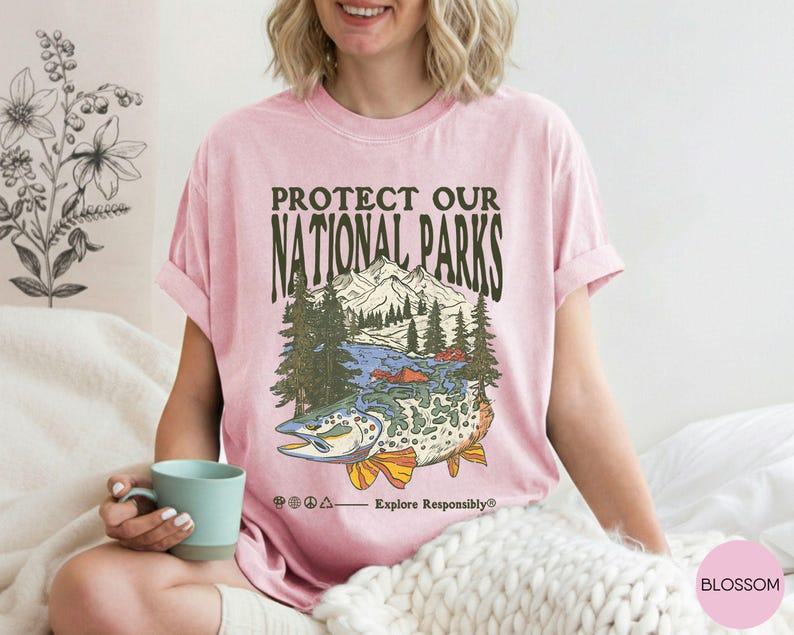 May include: Pink t-shirt with a graphic that says "Protect Our National Parks." The graphic features a mountain range, trees, a lake, and a fish. The text "Explore Responsibly" is also included.