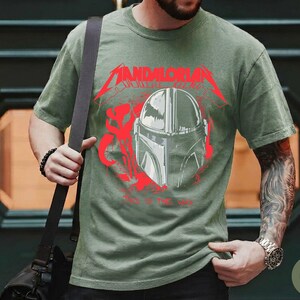 Op de afbeelding: Mosgroen t-shirt met een afbeelding van een Mandalorian helm en de woorden "Mandalorian" en "Way of the Jedi" in het rood. Het t-shirt heeft een ronde hals.