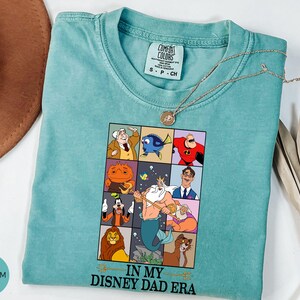 May include: Seafoam green Comfort Colors t-shirt with a graphic featuring Disney characters in a grid. The phrase "IN MY DISNEY DAD ERA" is printed below the grid. A gold necklace is draped across the shirt.