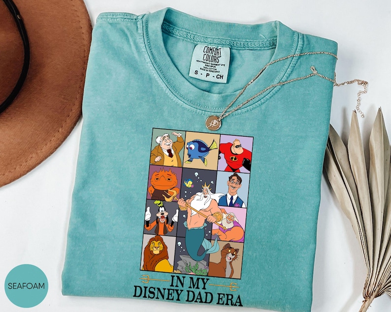 May include: Seafoam green Comfort Colors t-shirt with a graphic featuring Disney characters in a grid. The phrase "IN MY DISNEY DAD ERA" is printed below the grid. A gold necklace is draped across the shirt.