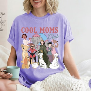 May include: A violet-colored t-shirt with the words "COOL MOMS Club" in a retro font. The shirt features cartoon illustrations of various characters. The shirt is made of a soft material and has short sleeves.