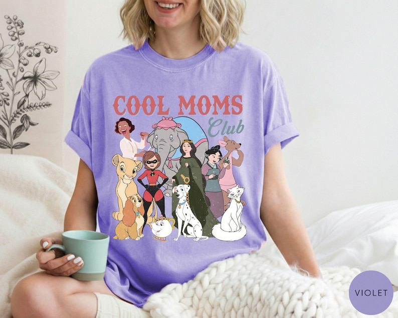 May include: A violet-colored t-shirt with the words "COOL MOMS Club" in a retro font. The shirt features cartoon illustrations of various characters. The shirt is made of a soft material and has short sleeves.