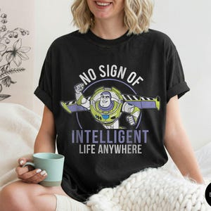 Puede incluir: Camiseta negra con un dise&ntilde;o de dibujos animados de Buzz Lightyear y el texto "NO SIGN OF INTELLIGENT LIFE ANYWHERE" en blanco y morado. Una prenda c&oacute;moda y casual.