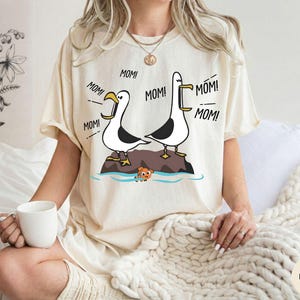 May include: Ivory-coloured t-shirt with a cartoon illustration of two seagulls on a rock, with the word "MOM!" repeated around them. A small orange fish is in the water below. The shirt has a relaxed fit.