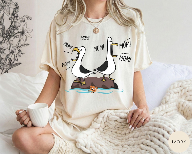 May include: Ivory-coloured t-shirt with a cartoon illustration of two seagulls on a rock, with the word "MOM!" repeated around them. A small orange fish is in the water below. The shirt has a relaxed fit.
