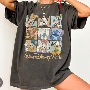 Może przedstawiać: Ciemnoszara koszulka z kolorowym wzorem w kratkę przedstawiającym postacie Disneya, w tym Myszka Miki, Myszka Minnie, Goofy, Kaczor Donald, Kaczka Daisy, Chip i Dale, Stitch i Pluto. Poniżej nadruk "Walt Disney World".