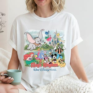 May include: A white short-sleeve t-shirt featuring a colorful graphic print of various Disney characters including Dumbo, Ariel, Mickey Mouse, and Goofy in front of a castle. The text Magic Kingdom and Walt Disney World is printed below the characters.