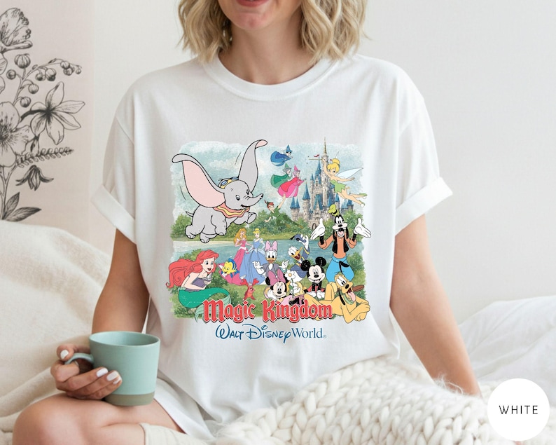 May include: A white short-sleeve t-shirt featuring a colorful graphic print of various Disney characters including Dumbo, Ariel, Mickey Mouse, and Goofy in front of a castle. The text Magic Kingdom and Walt Disney World is printed below the characters.