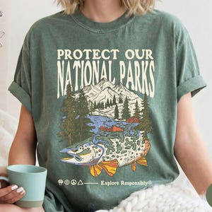May include: Moss green t-shirt with a graphic that reads "Protect Our National Parks." The graphic features a mountain range, trees, a river, and a fish. The shirt also has the text "Explore Responsibly."