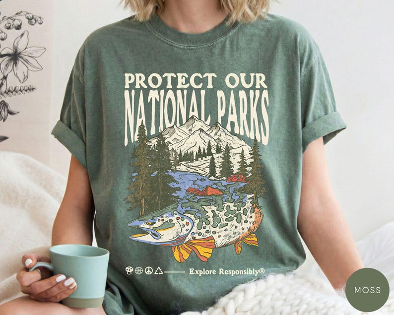 May include: Moss green t-shirt with a graphic that reads "Protect Our National Parks." The graphic features a mountain range, trees, a river, and a fish. The shirt also has the text "Explore Responsibly."
