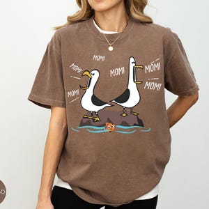 May include: Espresso-coloured t-shirt with a cartoon illustration of two seagulls on a rock, with the word "MOM!" coming from their beaks. A small orange fish is in the water below. The shirt has a relaxed fit.